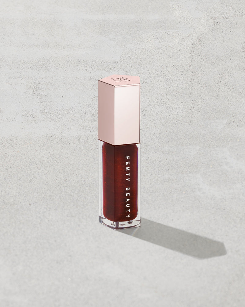 Fenty Beauty liquid lipstick in a deep berry red shade, housed in a clear bottle with a metallic rose gold cap, set against a textured light gray background.