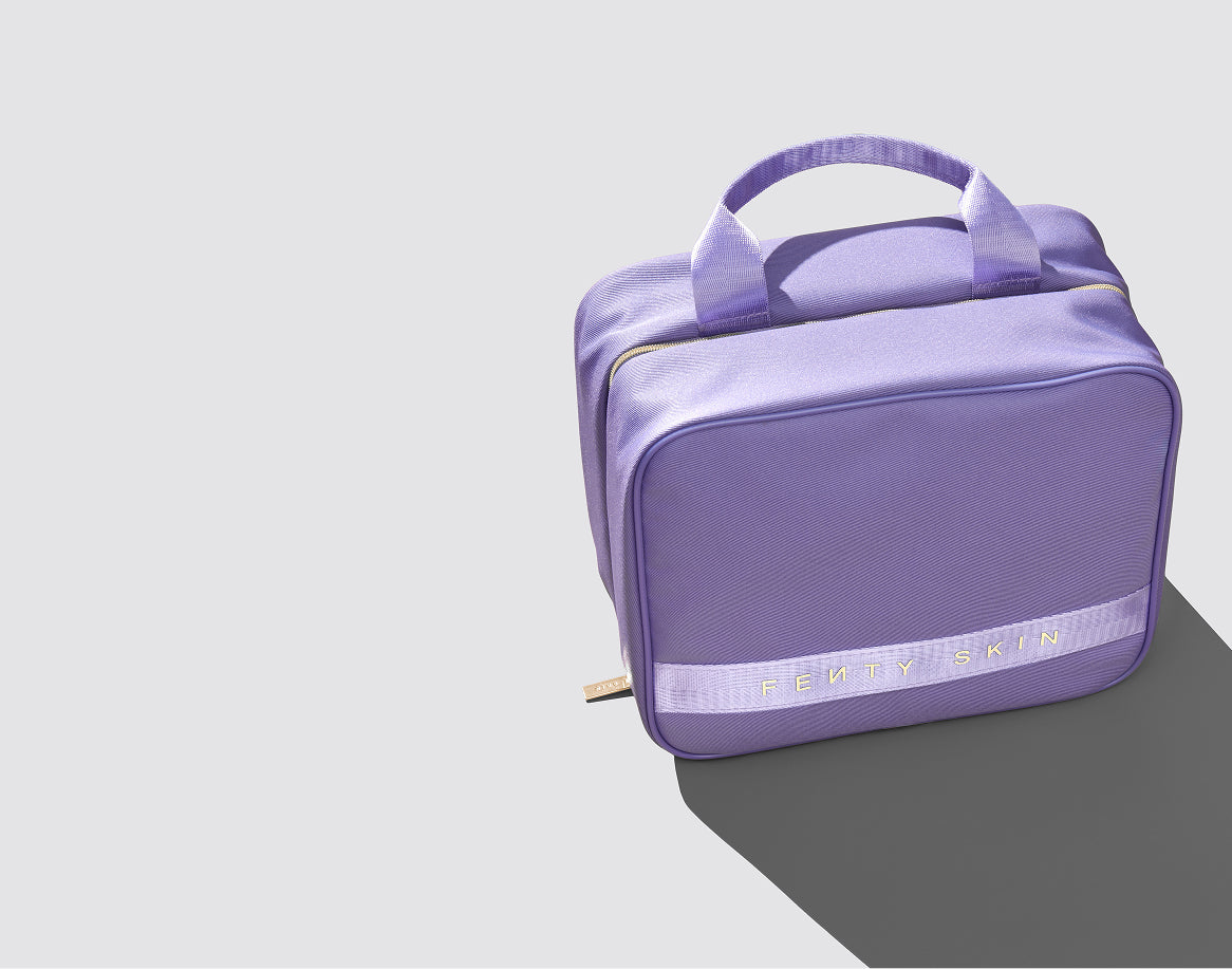 A lavender purple toiletry bag with dual top handles, a zippered top, and a horizontal branded band near the bottom, shown on a light gray background with a dark shadow.