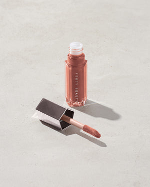 An open tube of Gloss Bomb Universal Lip Luminizer in the shade Fenty Glow on a grey background.