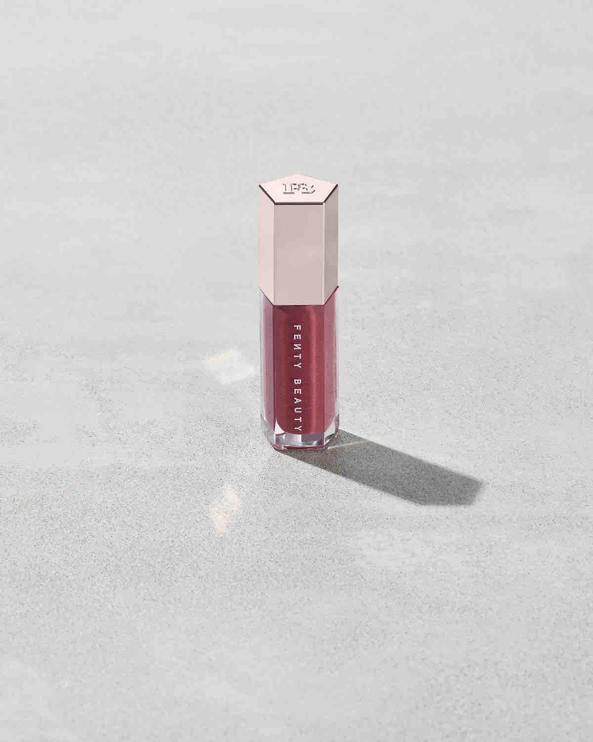 A closed tube of Gloss Bomb Universal Lip Luminizer in the shade RiRi on a grey background.