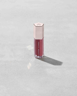A closed tube of Gloss Bomb Universal Lip Luminizer in the shade RiRi on a grey background.