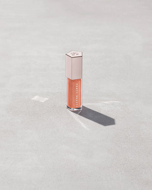 A closed tube of Gloss Bomb Universal Lip Luminizer in the shade Champ Stamp Fantasy on a grey background.