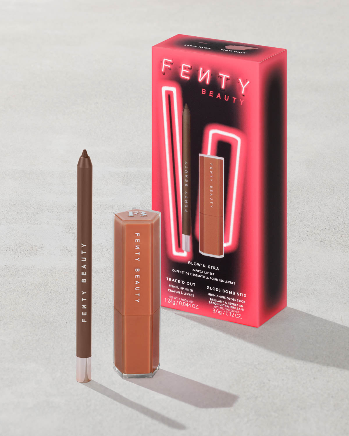 Fenty Beauty Glow N' Extra packaging with eyeliner and Gloss Bomb Stix with a gray concrete background