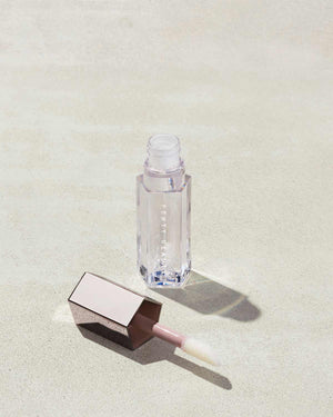 An open tube of Gloss Bomb Universal Lip Luminizer in the shade Glass Slipper on a grey background.