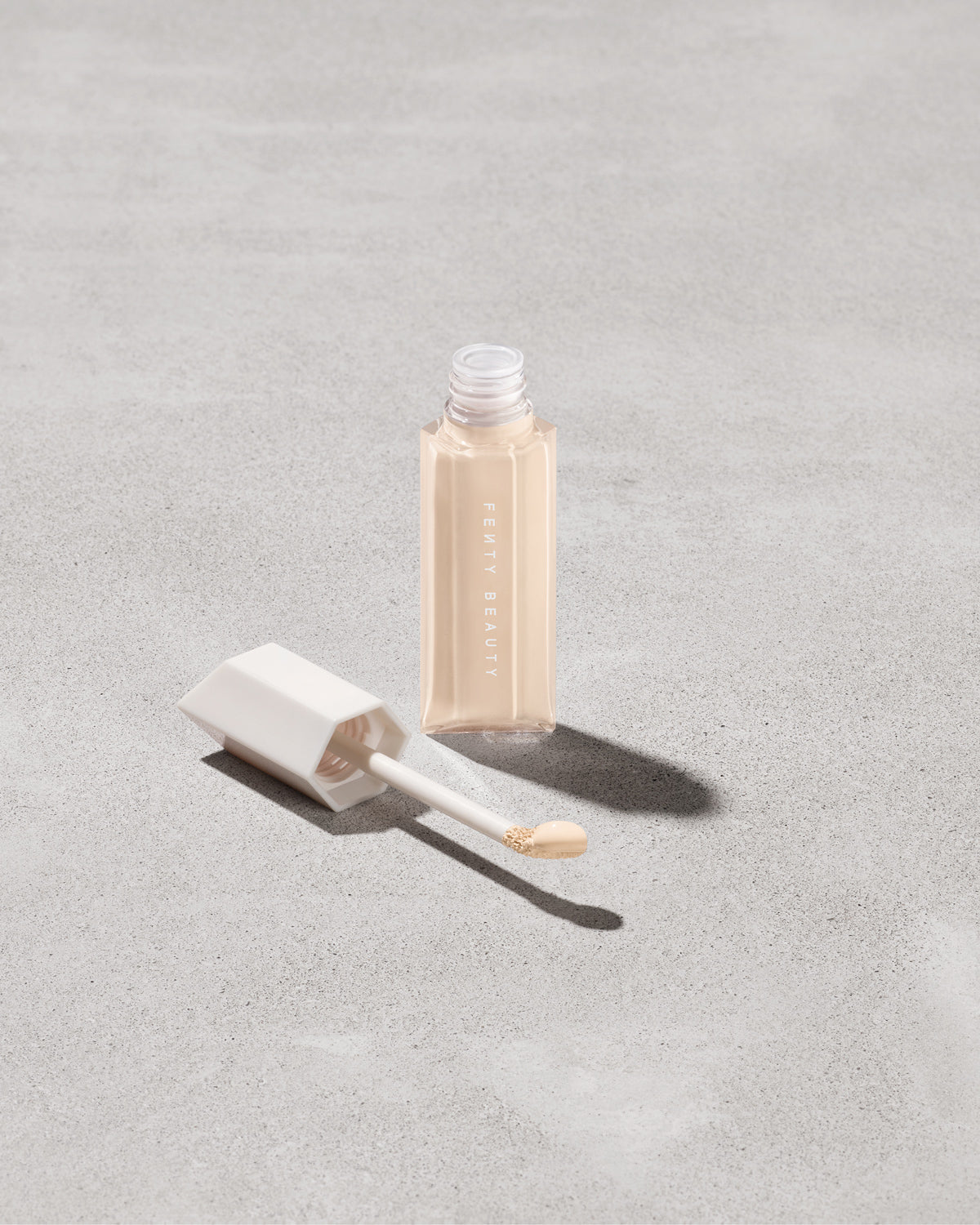 Open bottle of Fenty Beauty We're Even Hydrating Longwear Concealer with doe foot applicator in shade 120n on a concrete background.