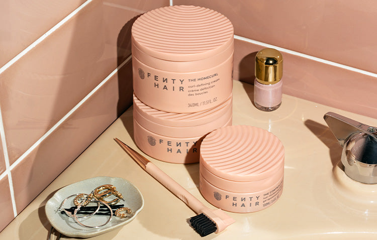 Product shot of Fenty Hair The Homecurl Curl-Defining Cream, The Controlling Type Edge Control Gel, and the Edge Control Stick stacked on the corner of a bathtub with blush pink tiles.