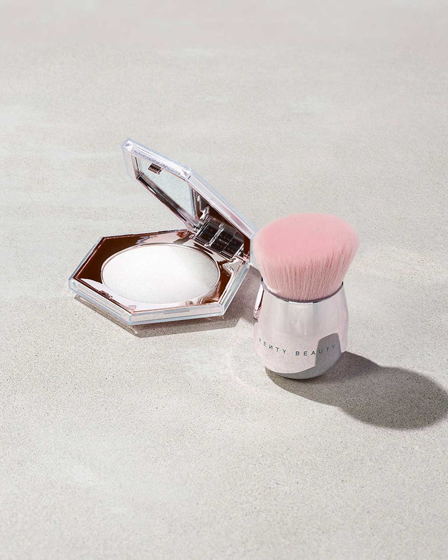 A fenty face and body kabuki brush stood next to an open powder container on a gray background