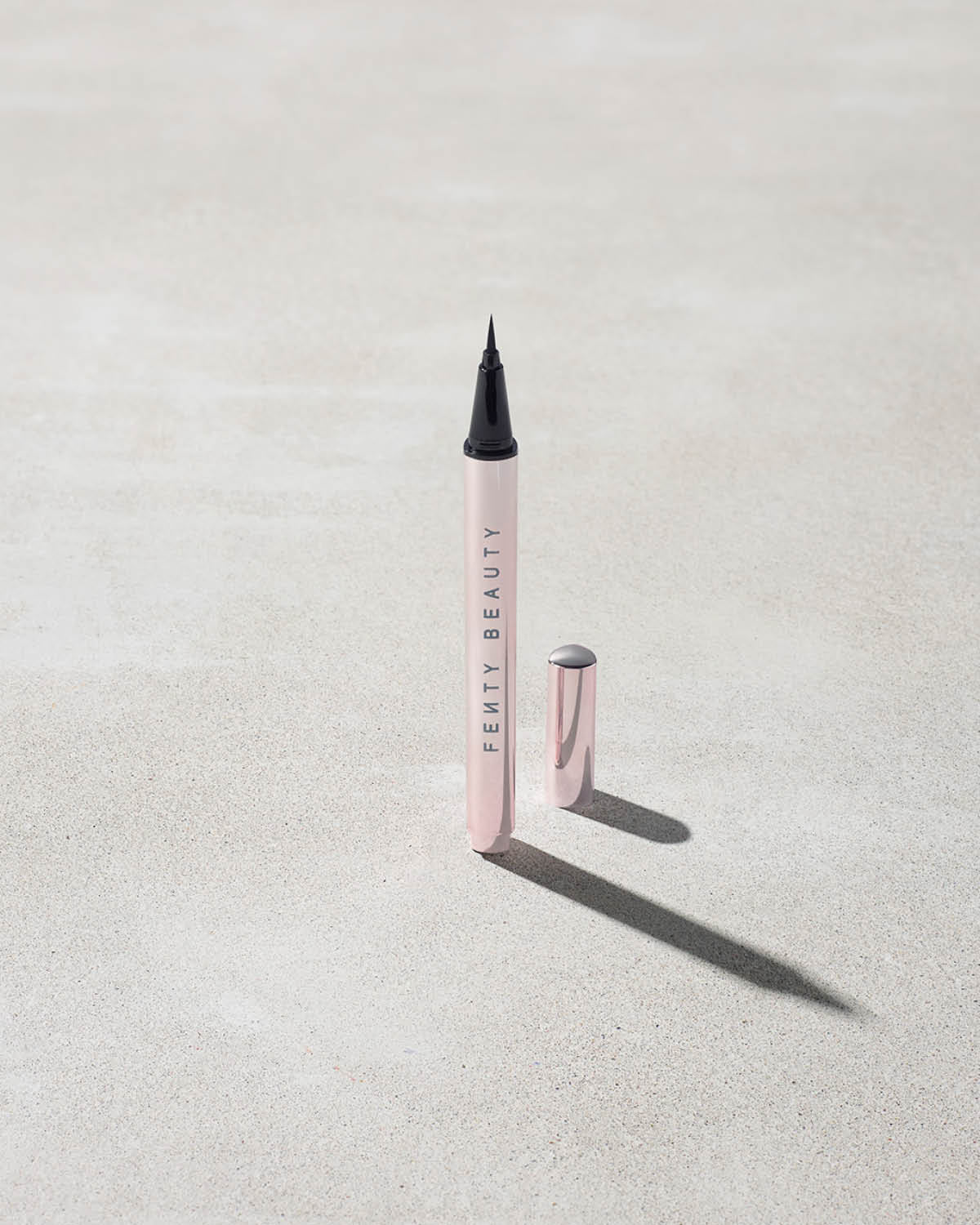 The Flyliner Longwear Liquid Eyeliner standing upright on a light grey concrete surface. The eyeliner is open, revealing its bold black tip, while its exterior features a sleek light metallic pink finish.